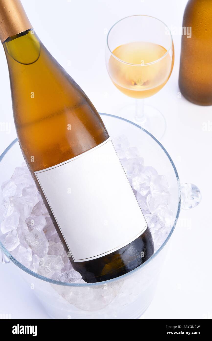 Primo piano di una bottiglia di vino bianco con etichetta vuota in un secchio di ghiaccio cristallino con un bicchiere e una bottiglia sullo sfondo . Foto Stock