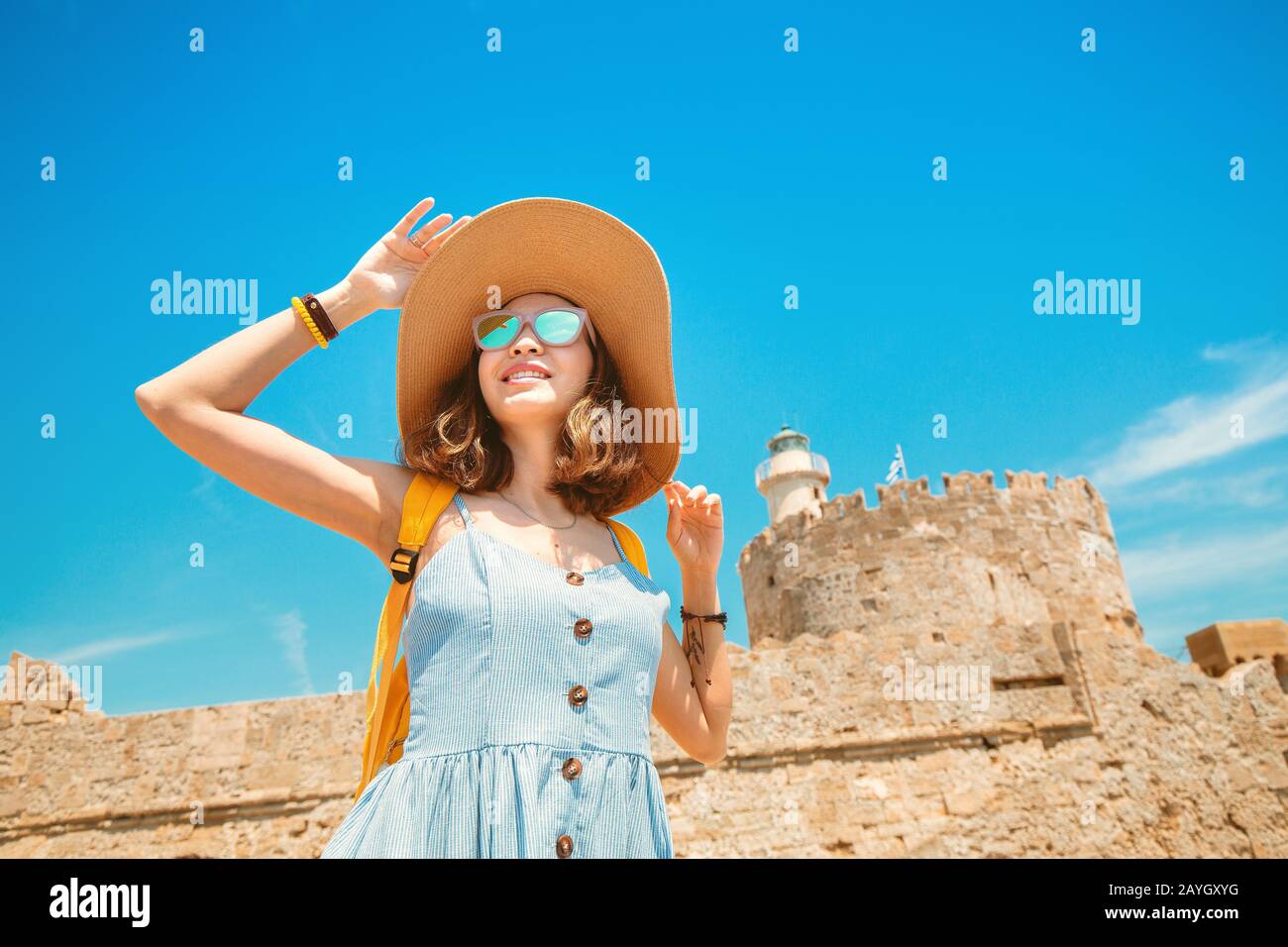 Felice donna turistica in vacanza posa con cappello e zaino di fronte alla vecchia fortezza in rovina e faro a Rodi, Grecia Foto Stock