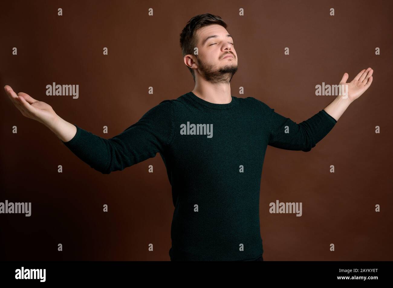 Giovane uomo con capelli castani vestito casualmente in una blusa verde che guarda in su e grazie con braccia aperte isolate su sfondo marrone Foto Stock