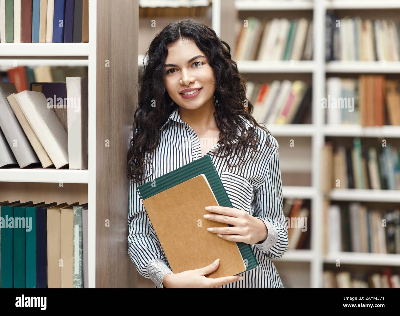 Donna latina sorridente che tiene libri di testo in biblioteca Foto Stock