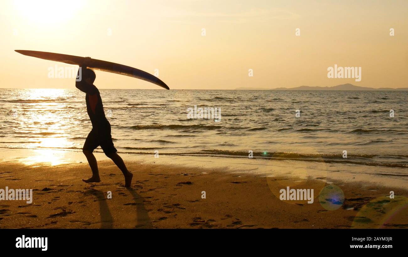 silhouette di uomo surf felice che corre con lunghe tavole da surf al tramonto su spiaggia tropicale. surfista sulla spiaggia in riva al mare al tramonto con bello Foto Stock