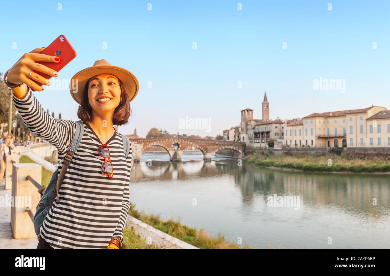 Sorridente donna viaggiatore che prende selfie sul suo smartphone sullo sfondo del Ponte pietra di Verona, in Italia Foto Stock