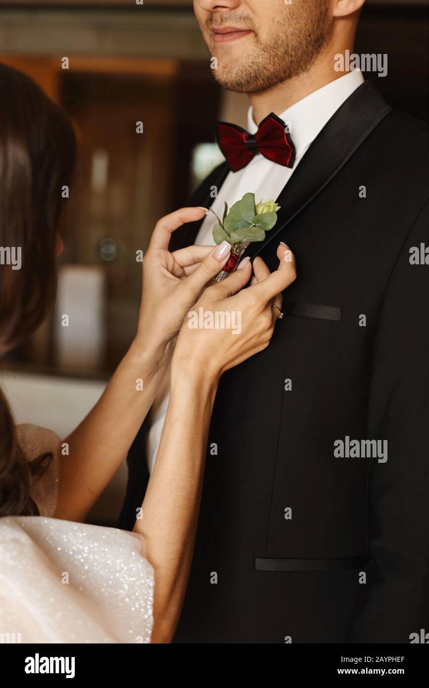 Vista ravvicinata delle mani femminili che fissano il boutonniere sulla giacca di un bell'uomo in un tuxedo e cravatta di prua. Ritratto a metà faccia di un uomo elegante in un Foto Stock