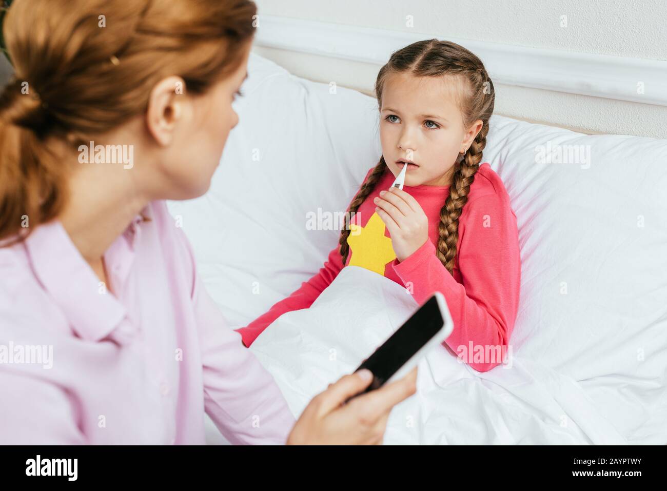 figlia ammalata che misura la febbre con il termometro mentre la madre che ha consultazione in linea sullo smartphone Foto Stock