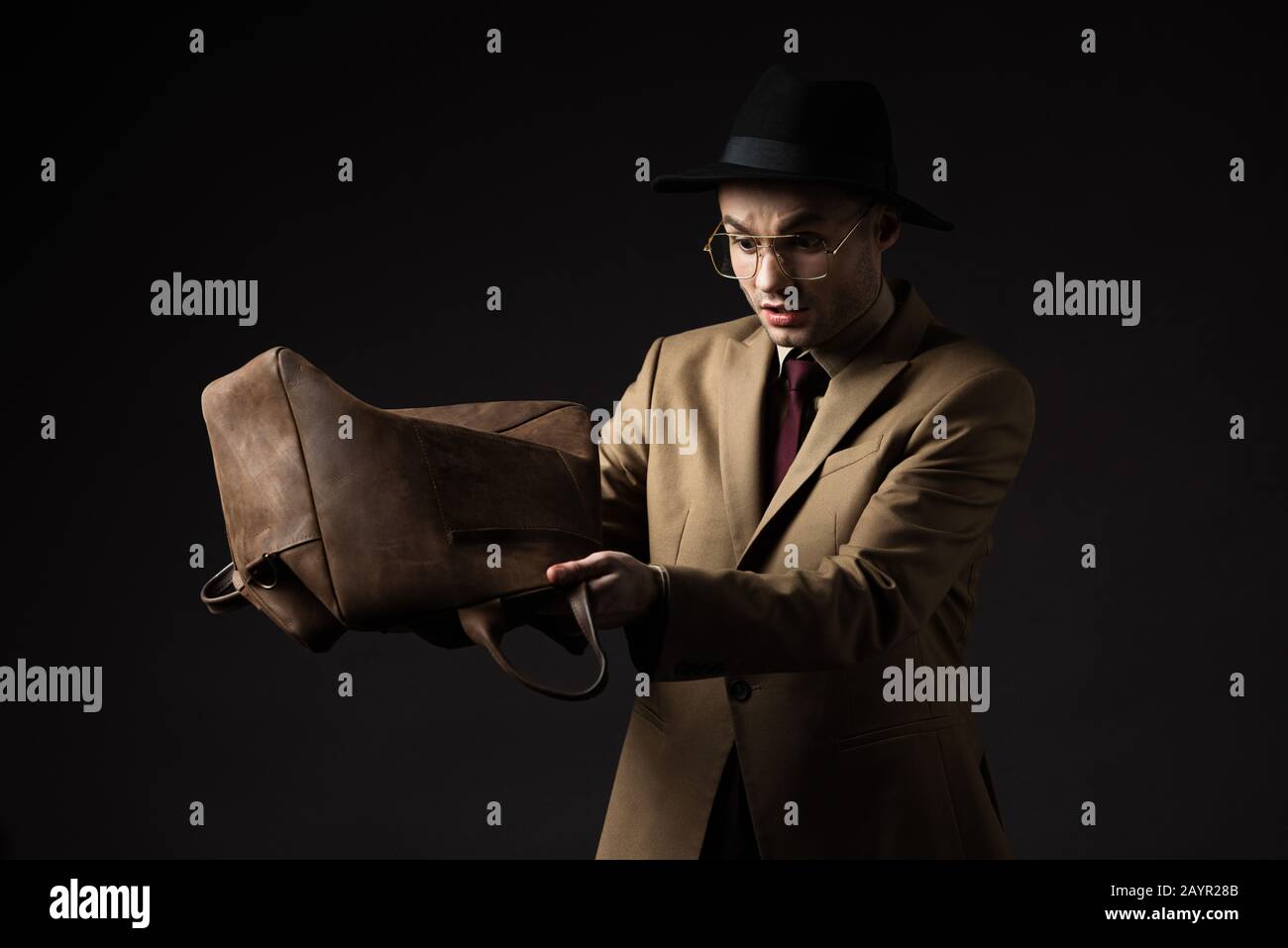 uomo elegante e stressato in abito beige, cappello e occhiali che scuotono fuori borsa di pelle marrone isolato su nero Foto Stock