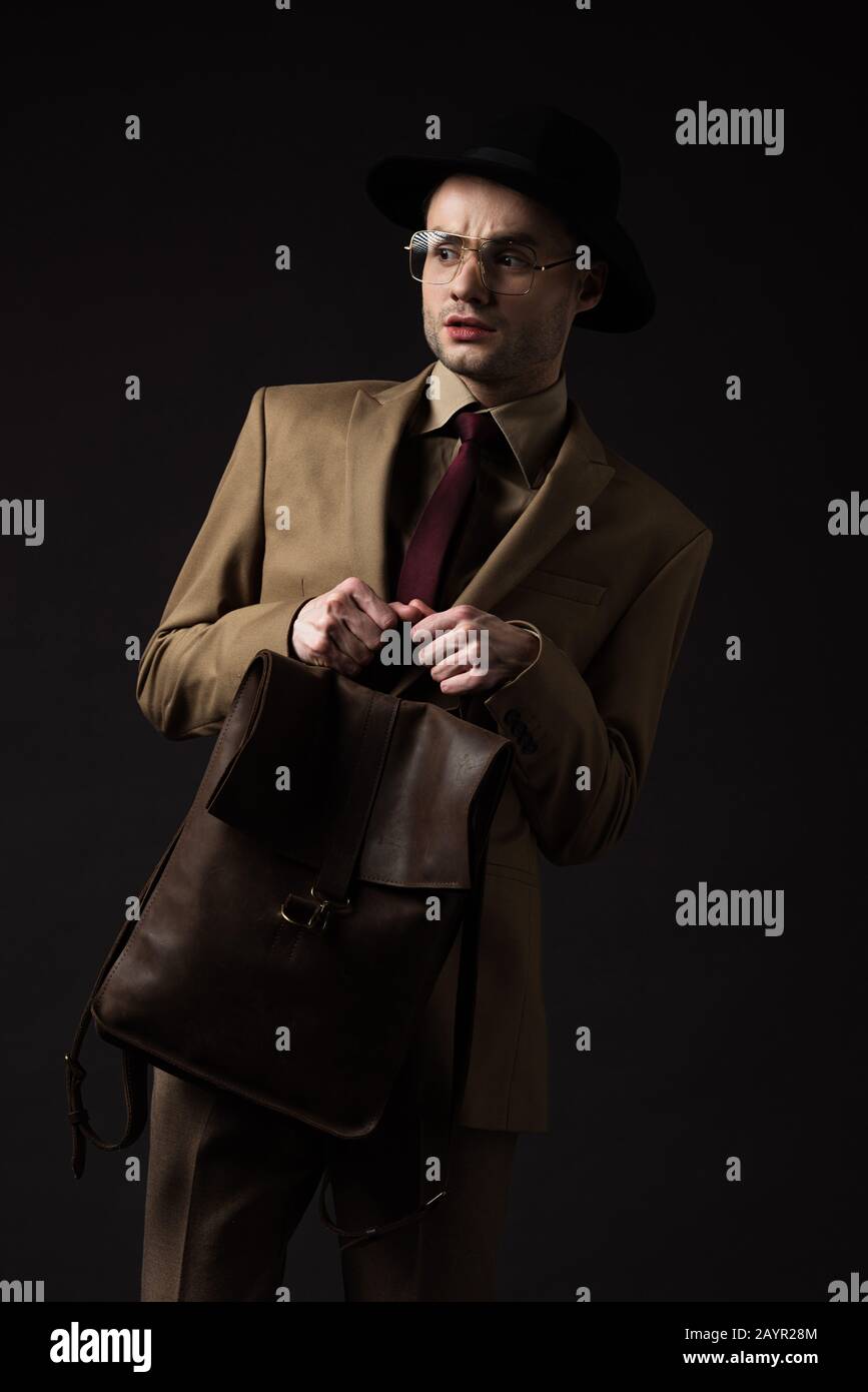 uomo elegante preoccupato in abito beige, cappello e occhiali che tengono zaino in pelle marrone isolato su nero Foto Stock