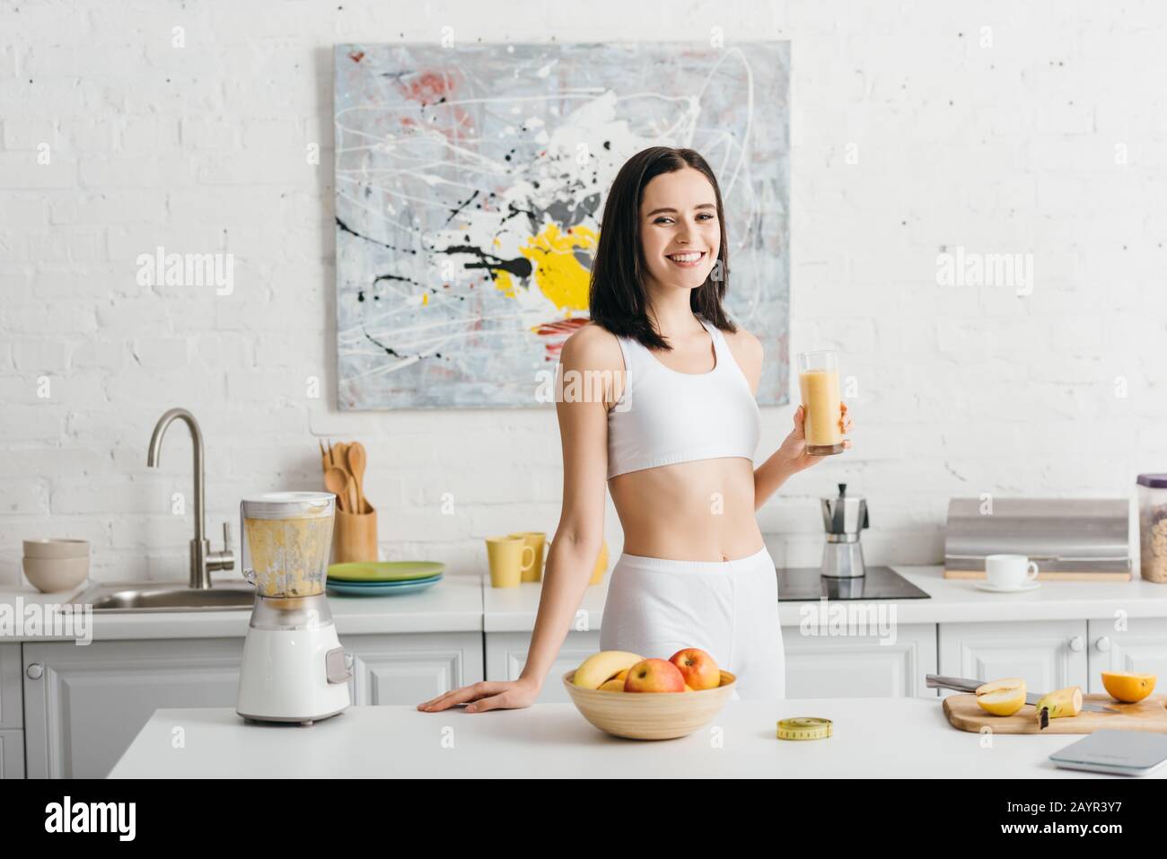 Attraente sportswoman sorridente alla macchina fotografica mentre tiene il vetro del frullato in cucina, dieta di conteggio delle calorie Foto Stock