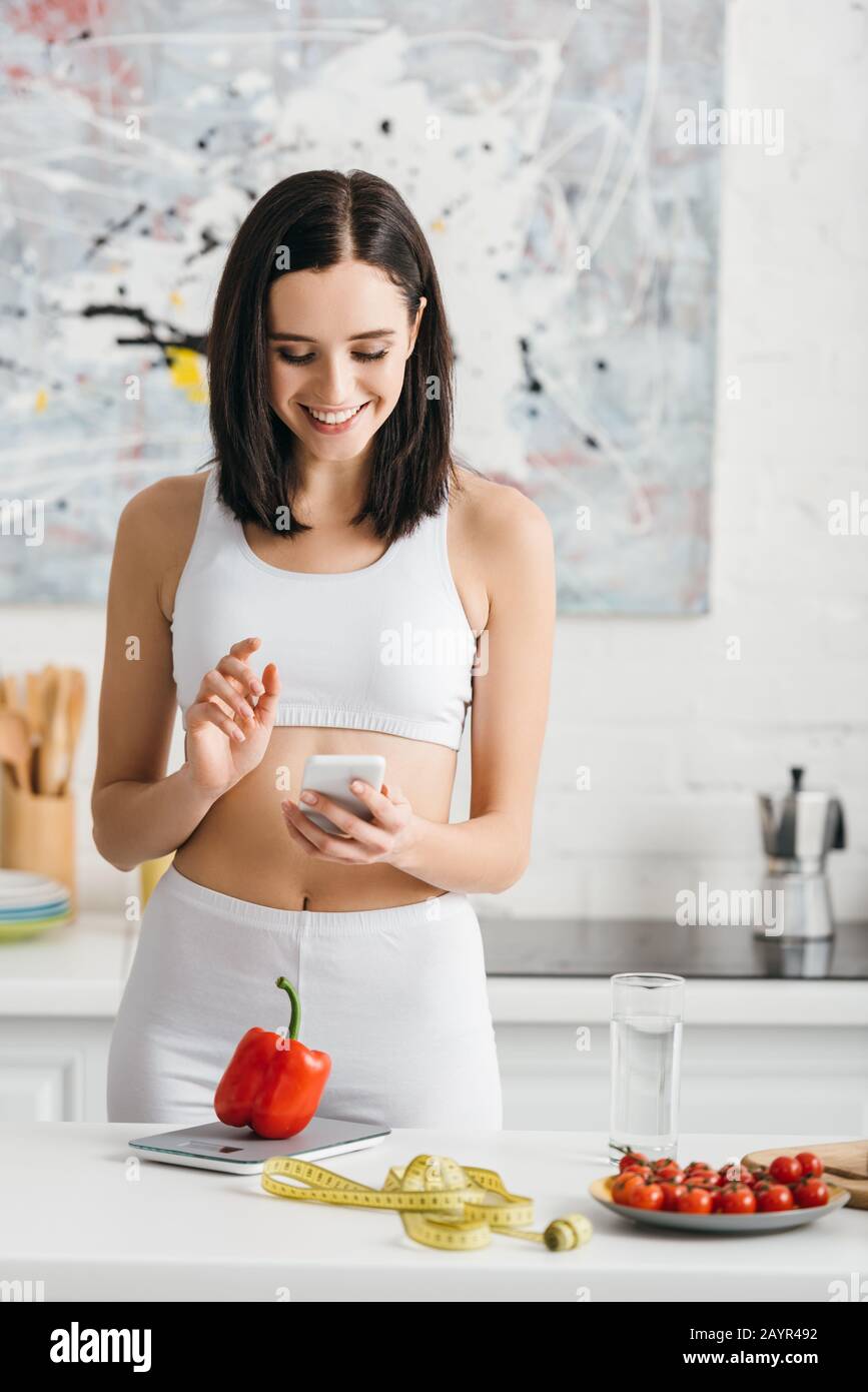 Sportswoman sorridente usando lo smartphone vicino a metro nastro, verdure e scale sul tavolo da cucina, dieta di conteggio delle calorie Foto Stock