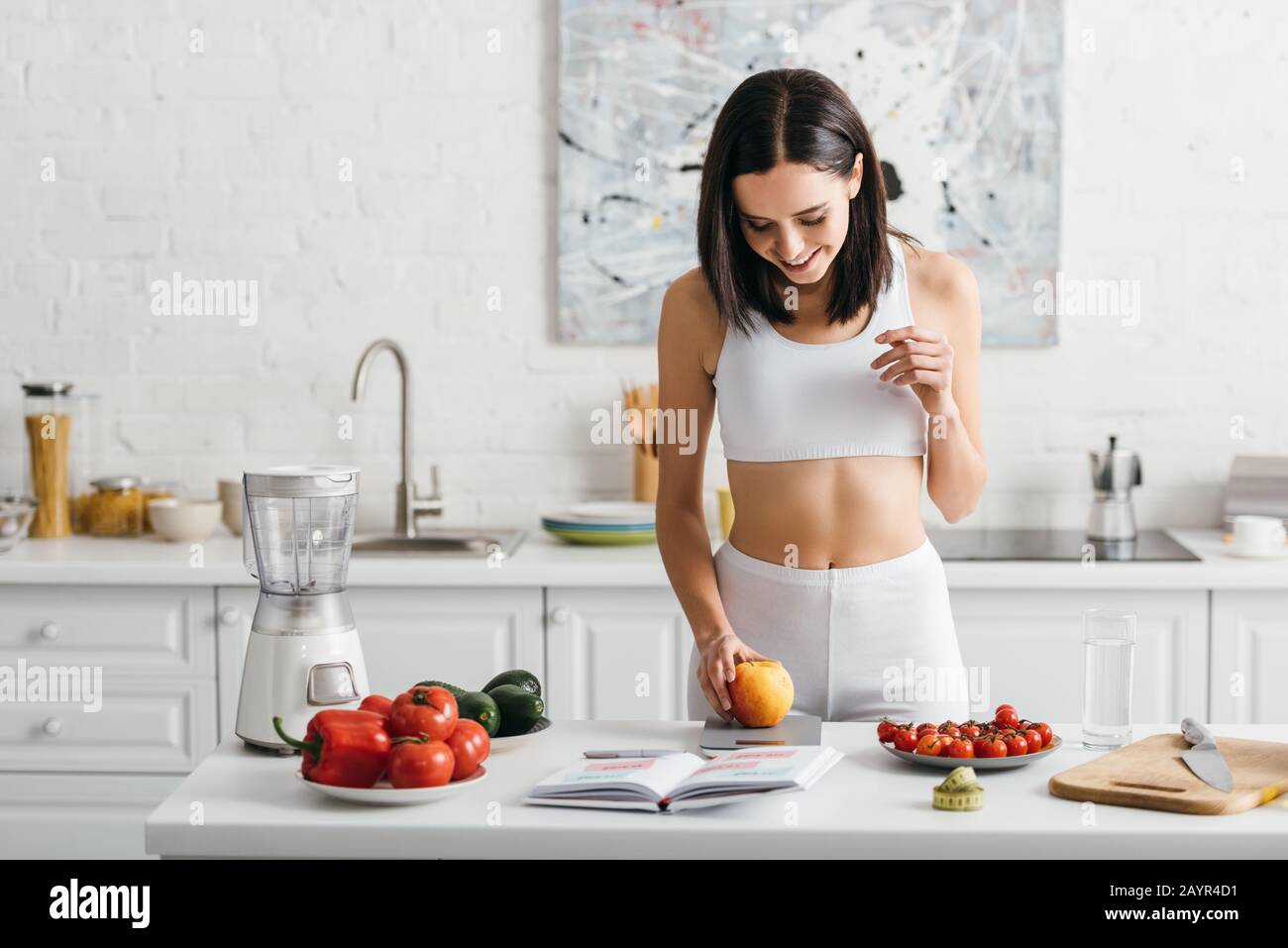 Sportswoman sorridente che pesano la mela vicino al taccuino e che misura il nastro sul tavolo della cucina, dieta di conteggio delle calorie Foto Stock