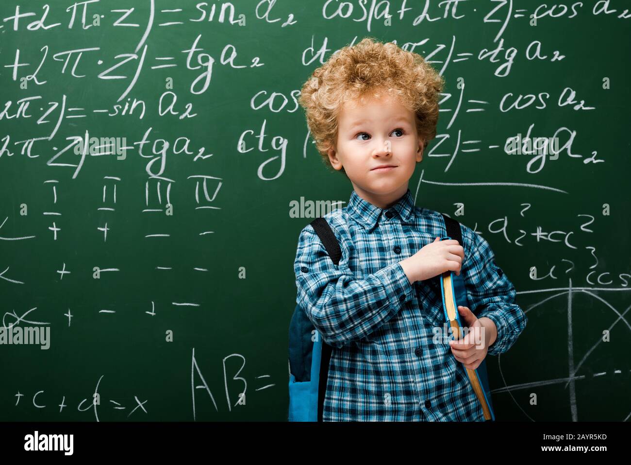 bambino intelligente in piedi con libro vicino lavagna con formule matematiche Foto Stock