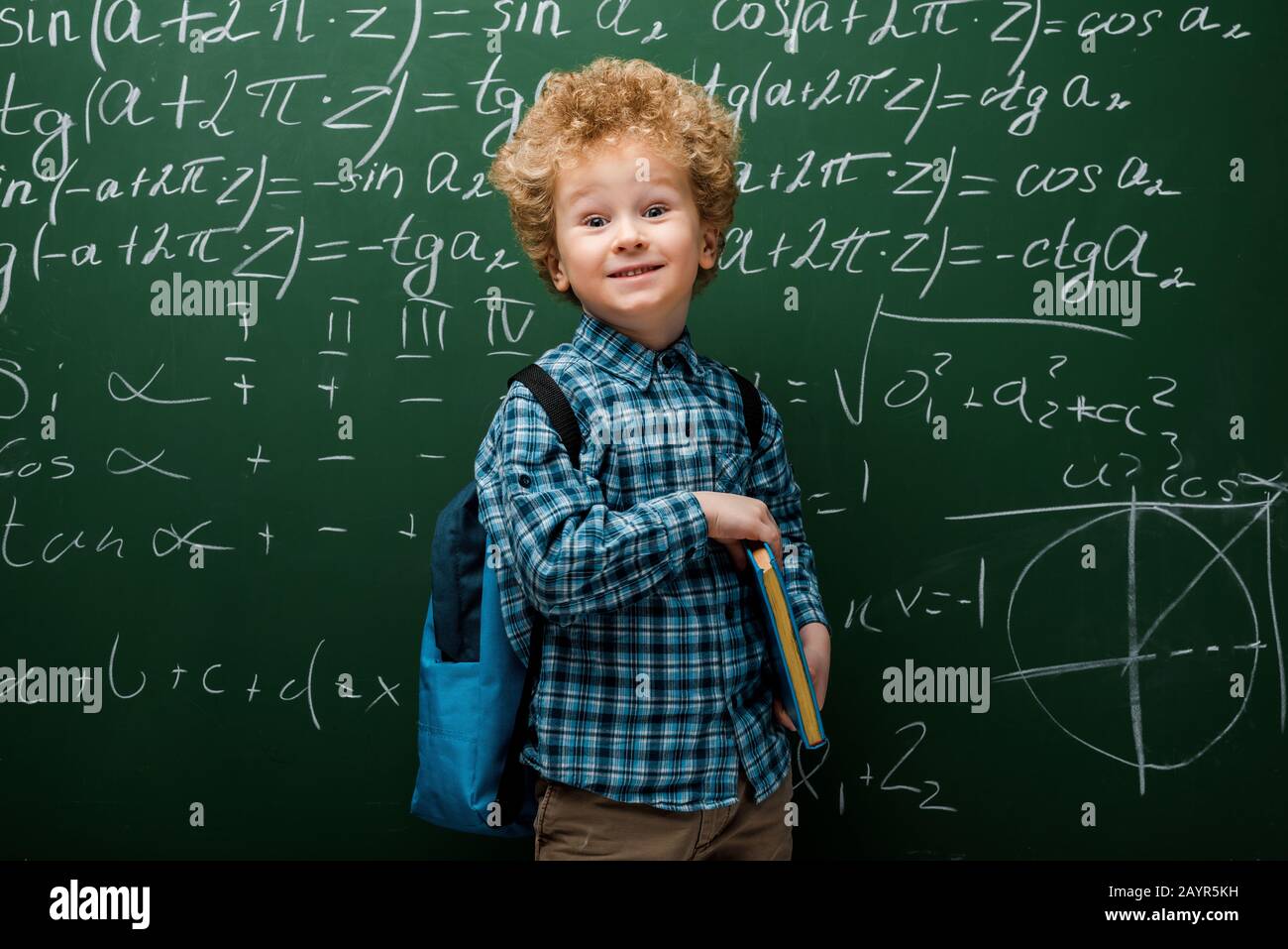 felice bambino che tiene libro vicino a lavagna con formule matematiche Foto Stock