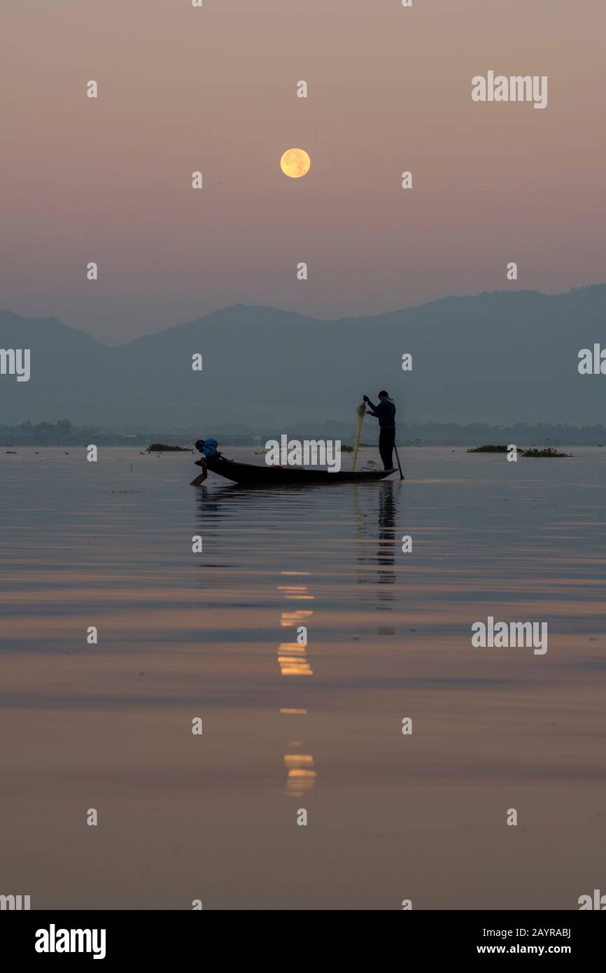 Prima dell'alba sotto una luna piena un pescatore di canottaggio della gamba nella sua barca sta tirando nella sua rete di pesca ha regolato il giorno prima sul lago di Inle in Myanmar. Foto Stock