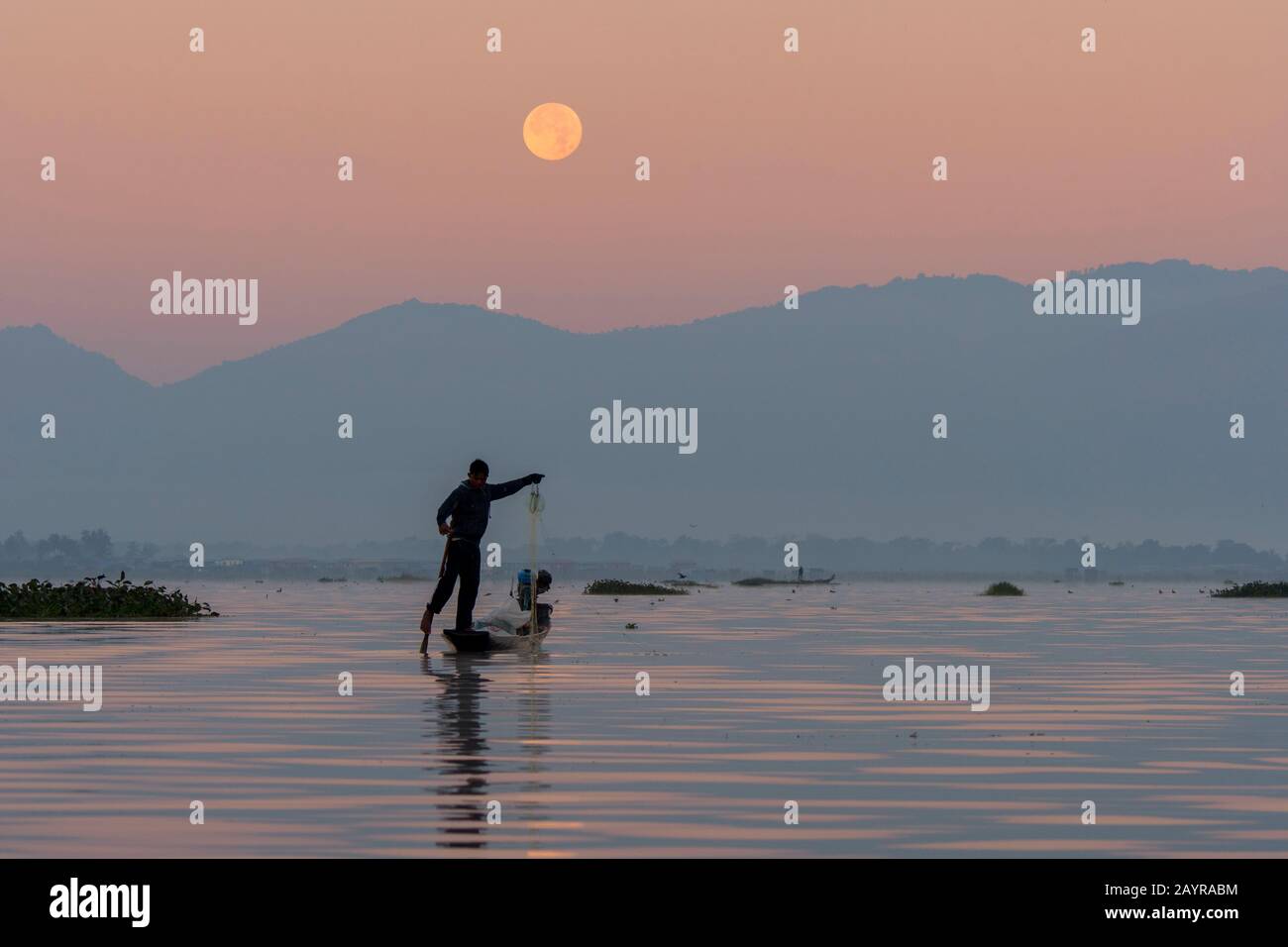 Prima dell'alba sotto una luna piena un pescatore di canottaggio della gamba nella sua barca sta tirando nella sua rete di pesca ha regolato il giorno prima sul lago di Inle in Myanmar. Foto Stock