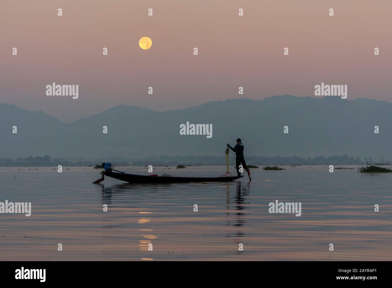 Prima dell'alba sotto una luna piena un pescatore di canottaggio della gamba nella sua barca sta tirando nella sua rete di pesca ha regolato il giorno prima sul lago di Inle in Myanmar. Foto Stock