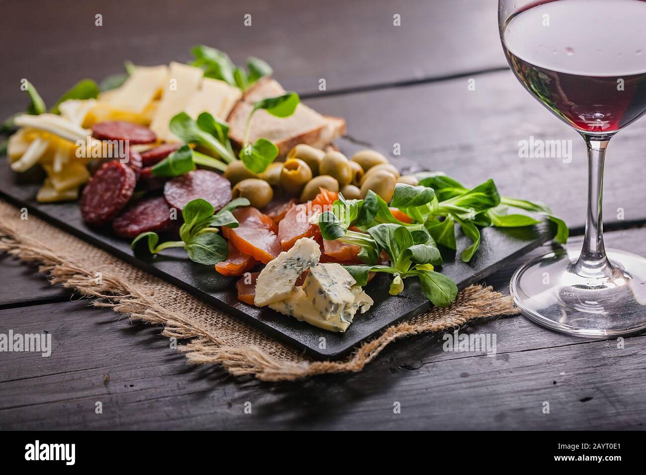 Spuntini mediterranei deliziosi e gustosi. Formaggi assortiti, salsicce, carne affumicata, lattuga, pane, olive, bicchiere di vino rosso e su un piatto piano scuro Foto Stock