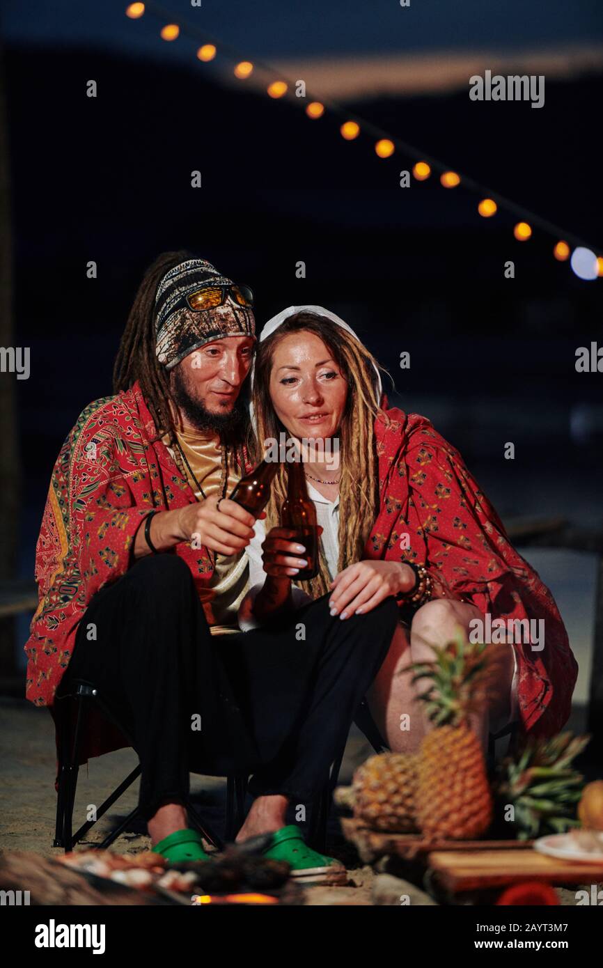 Una coppia copre con coperta e beve birra in bottiglia quando si trascorre la notte al caminetto sulla spiaggia Foto Stock