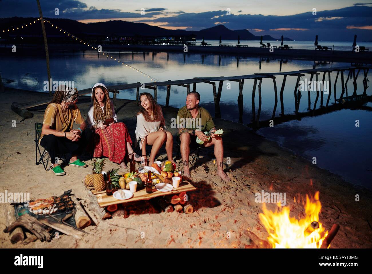 Gruppo di amici che riposano sulla spiaggia di notte, giocando ukulele e cibo di esting da camino Foto Stock
