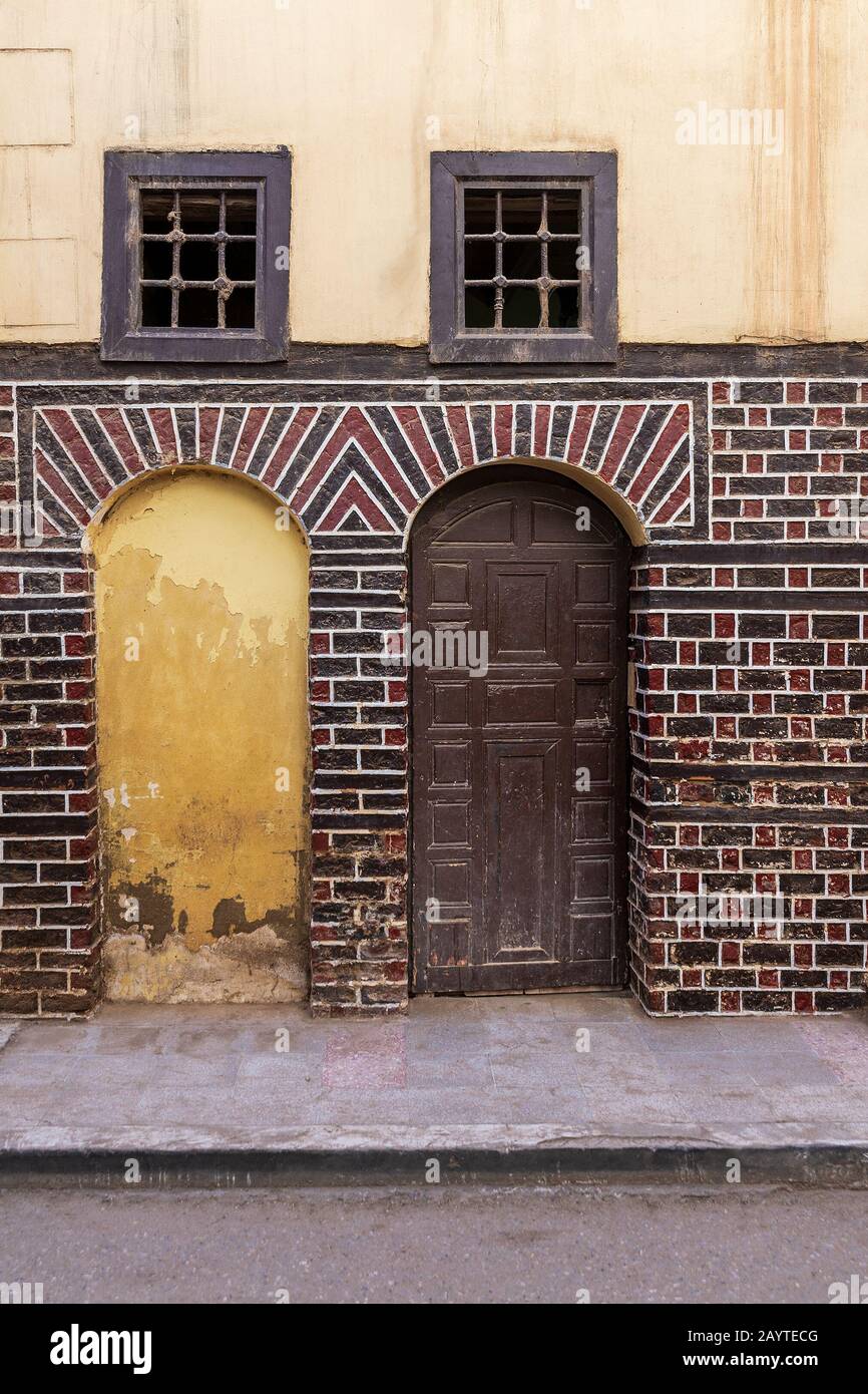 Porta in pietra bloccata, porta a volta decorata in legno, e due piccole finestre con barre in ferro arrugginito su mattoni neri e rossi muro su strada lastricata Foto Stock