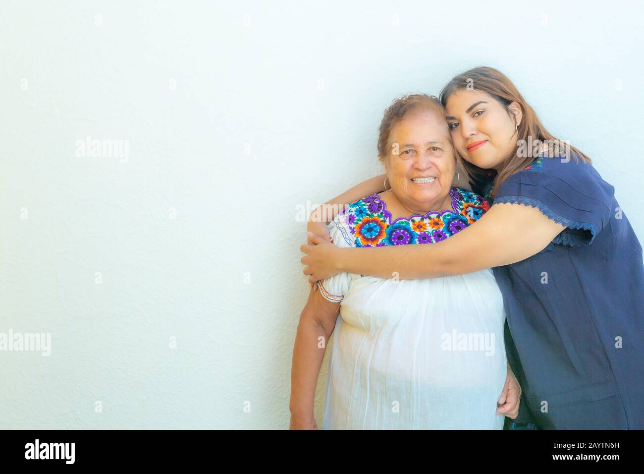 Nipote abbracciando sua nonna, due donne messicane sorridenti su sfondo bianco, spazio per il testo Foto Stock
