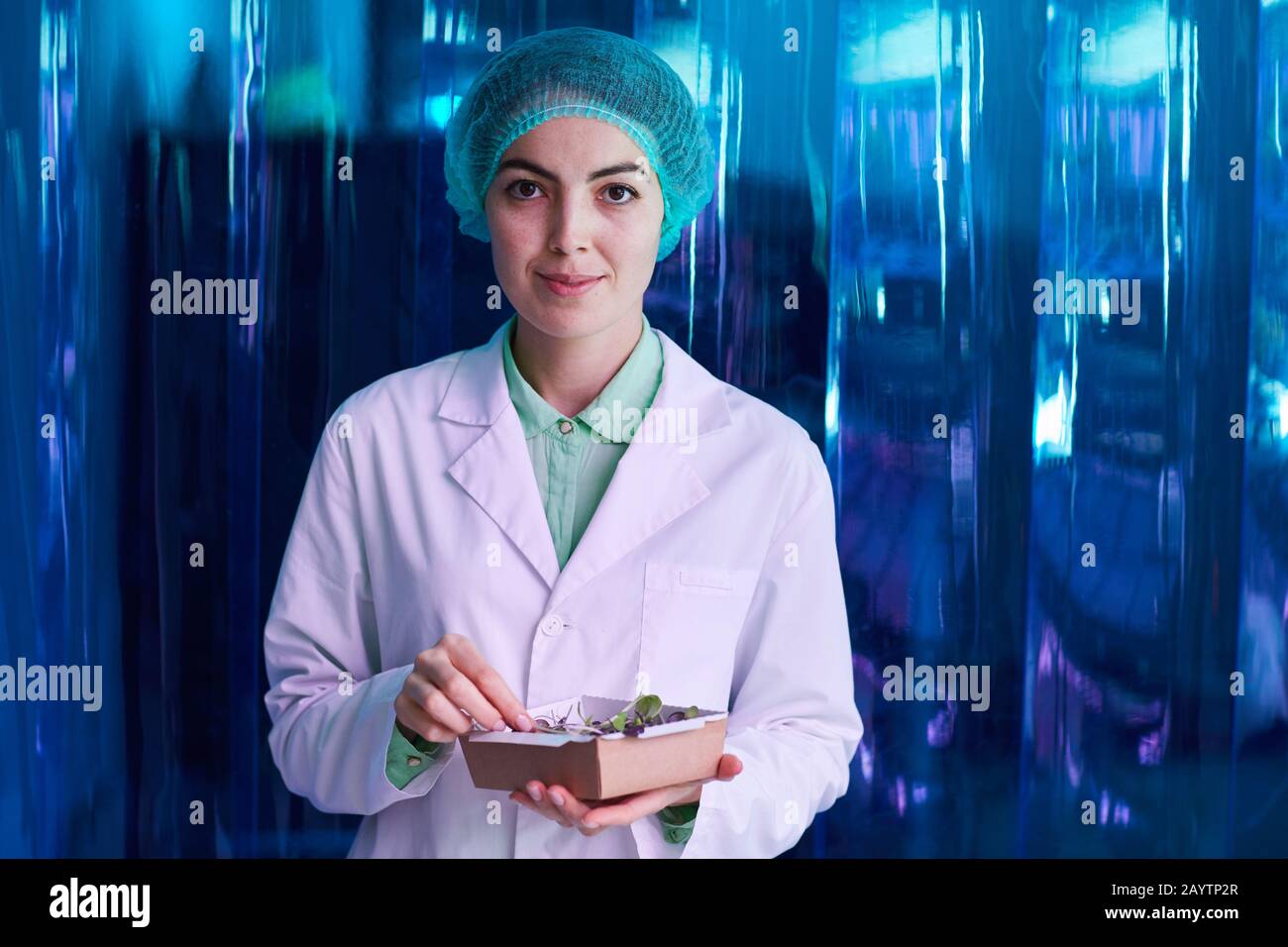 Girovita ritratto di sorridente scienziato femminile che tiene scatola di piante verdi mentre si posa contro la parete di plastica blu, copia spazio Foto Stock