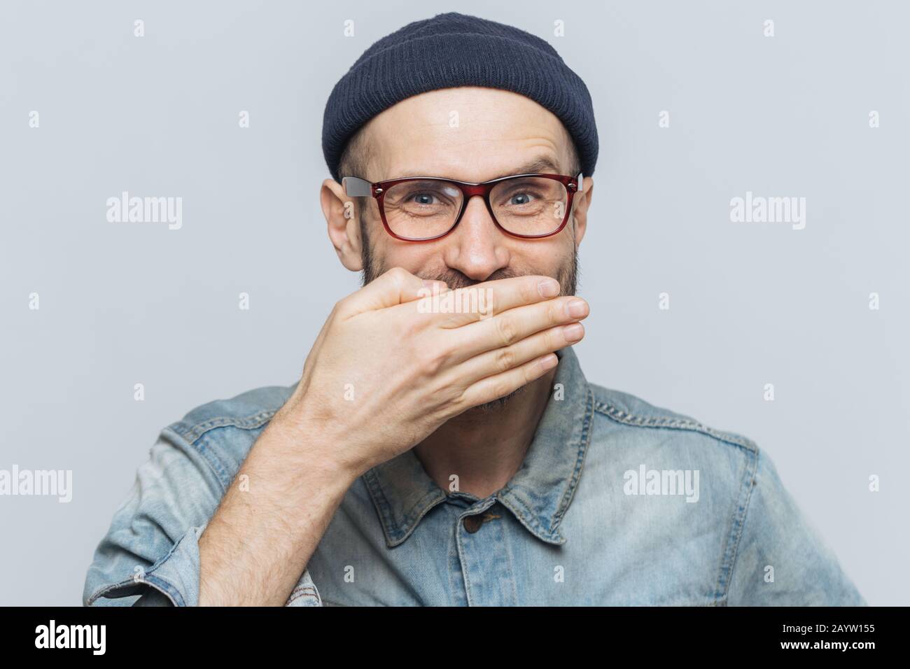Funny Man con look accattivante giggles o ride con gioia, copre la bocca con la mano, cerca di fermare i sentimenti, sente aneddoto da amico, indossa gli occhiali, Foto Stock
