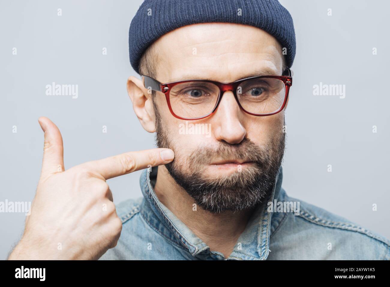 Immagine ravvicinata di barba lunga maschio con folta barba e baffi si brucia le guance e indica con il dito anteriore, essendo concentrata su qualcosa, isolato ov Foto Stock