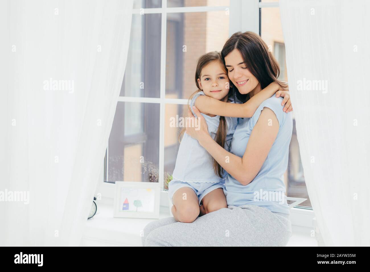 Grazioso piccolo bambino con un aspetto gradevole abbraccia la madre, esprime amore e buona sensazione o atteggiamento, sedersi sul davanzale, godetevi atmos domestico Foto Stock