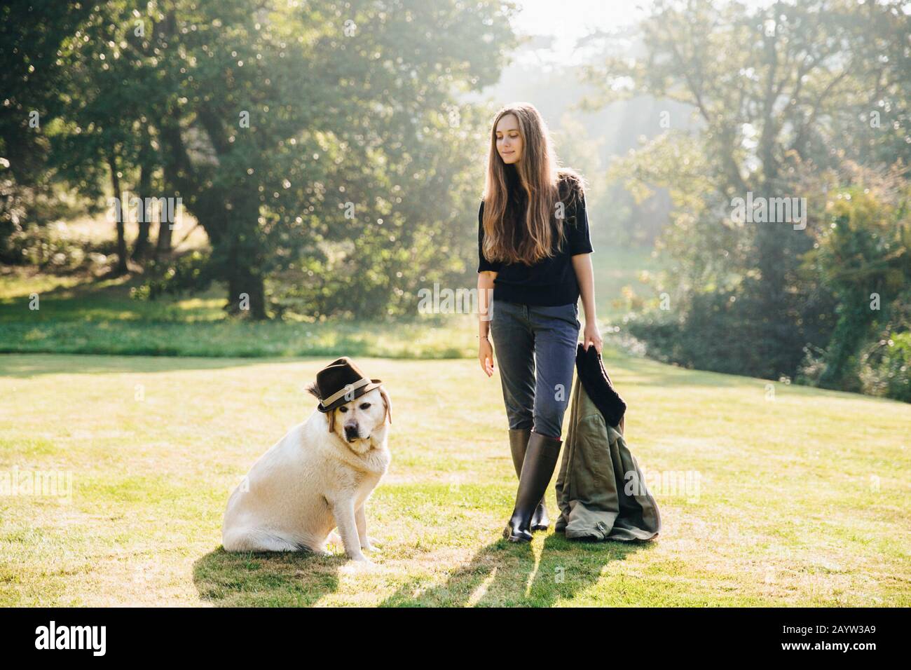 Bella giovane donna in piedi sull'erba, tenendo parka e guardando il cane divertente in cappello. Foto Stock