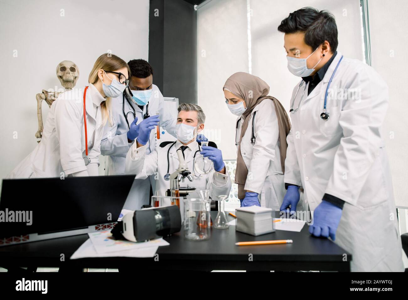 Gruppo di giovani scienziati multietnici della farmacia o della genetica in camici da laboratorio, guanti e maschere che lavorano insieme a microscopio, matracci e provette. Maschio Foto Stock