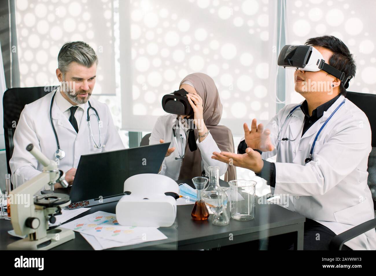 Due studenti di medicina o giovani scienziati che indossano occhiali di protezione per la realtà virtuale, studiano biochemistia o farmacologia, e il loro insegnante di sesso maschile che li guarda e fa Foto Stock