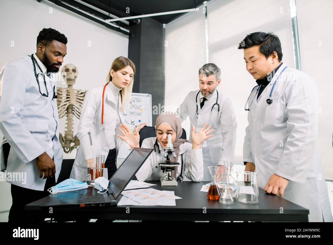 Gli scienziati della vita che ricercano in laboratorio. La giovane scienziato mussulmano femmina microscoping e gesturante allegramente mentre i suoi colleghi sono in piedi e. Foto Stock