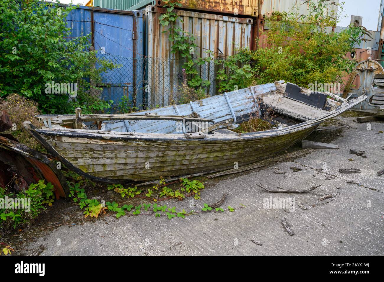 Clinker costruito barca di pesca di salmone chiamato Ulla quasi rotted via, posando in un cortile accanto ad alcuni contenitori di deposito. Foto Stock