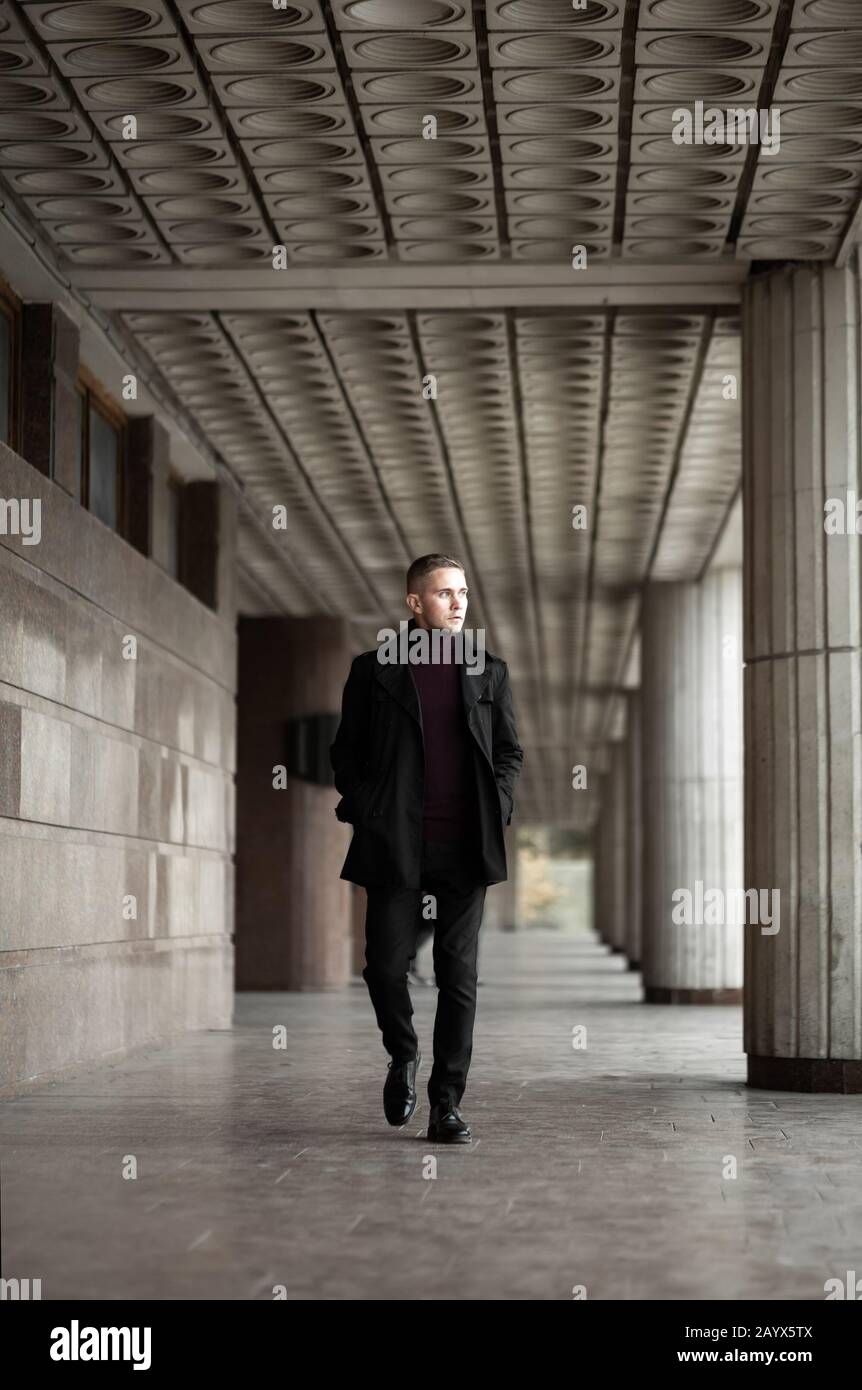 Uomo elegante in trench nero e passeggiate con il collo di polo presso l'edificio modernista Foto Stock