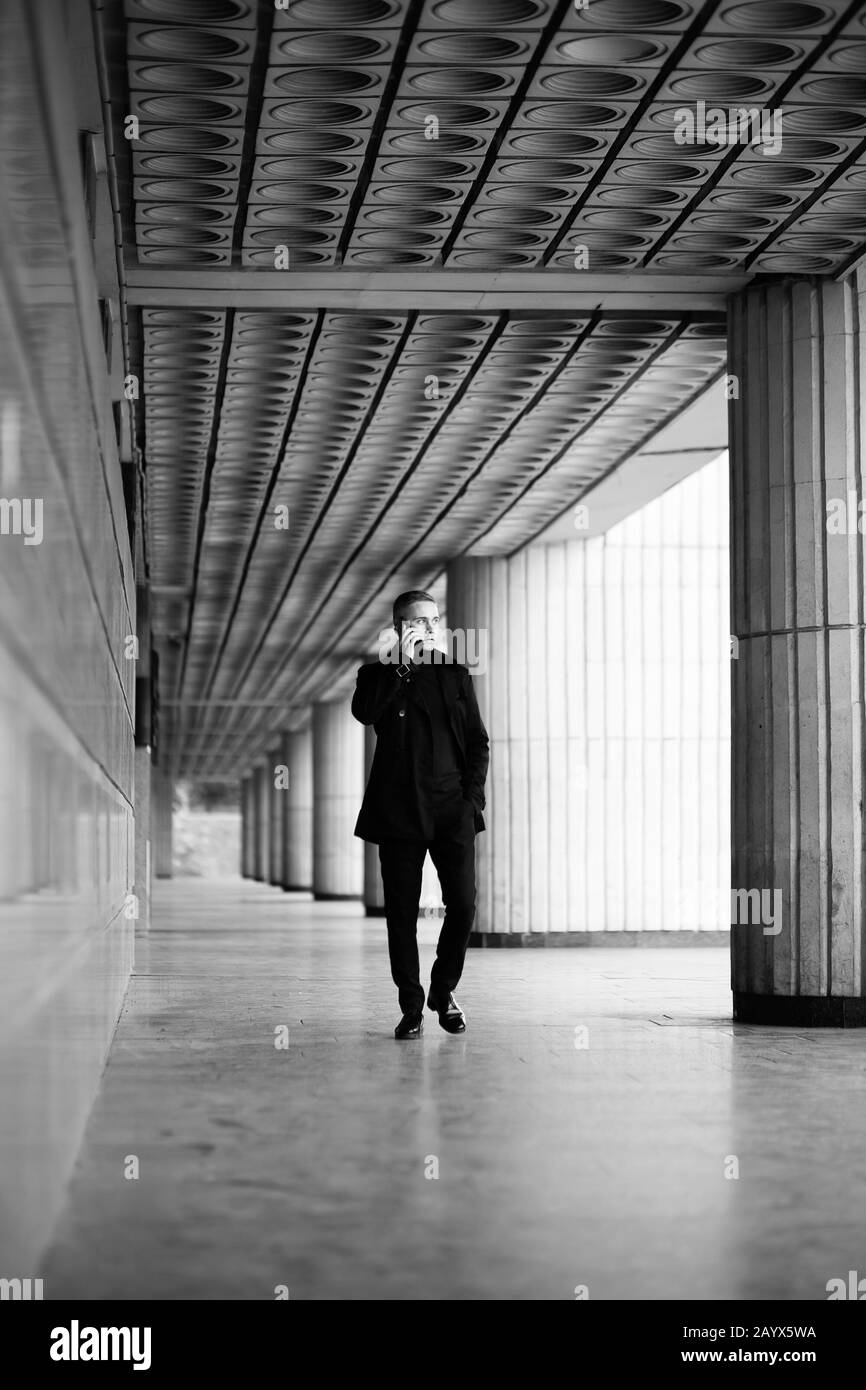Uomo elegante in trench nero e polo collo passeggiate presso l'edificio modernista e parlare su un telefono cellulare Foto Stock