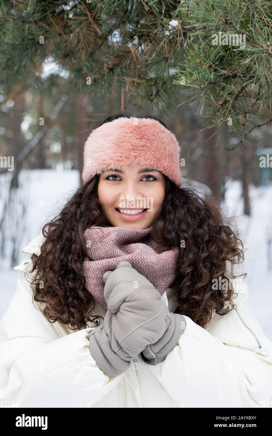Verticale medio inverno ritratto di attraente giovane donna con capelli ricci scuri che riscaldano le mani guardando la fotocamera sorridente Foto Stock