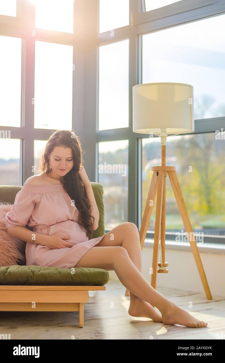 Giovane donna incinta che gode mattina soleggiata vicino alla grande finestra in casa Foto Stock