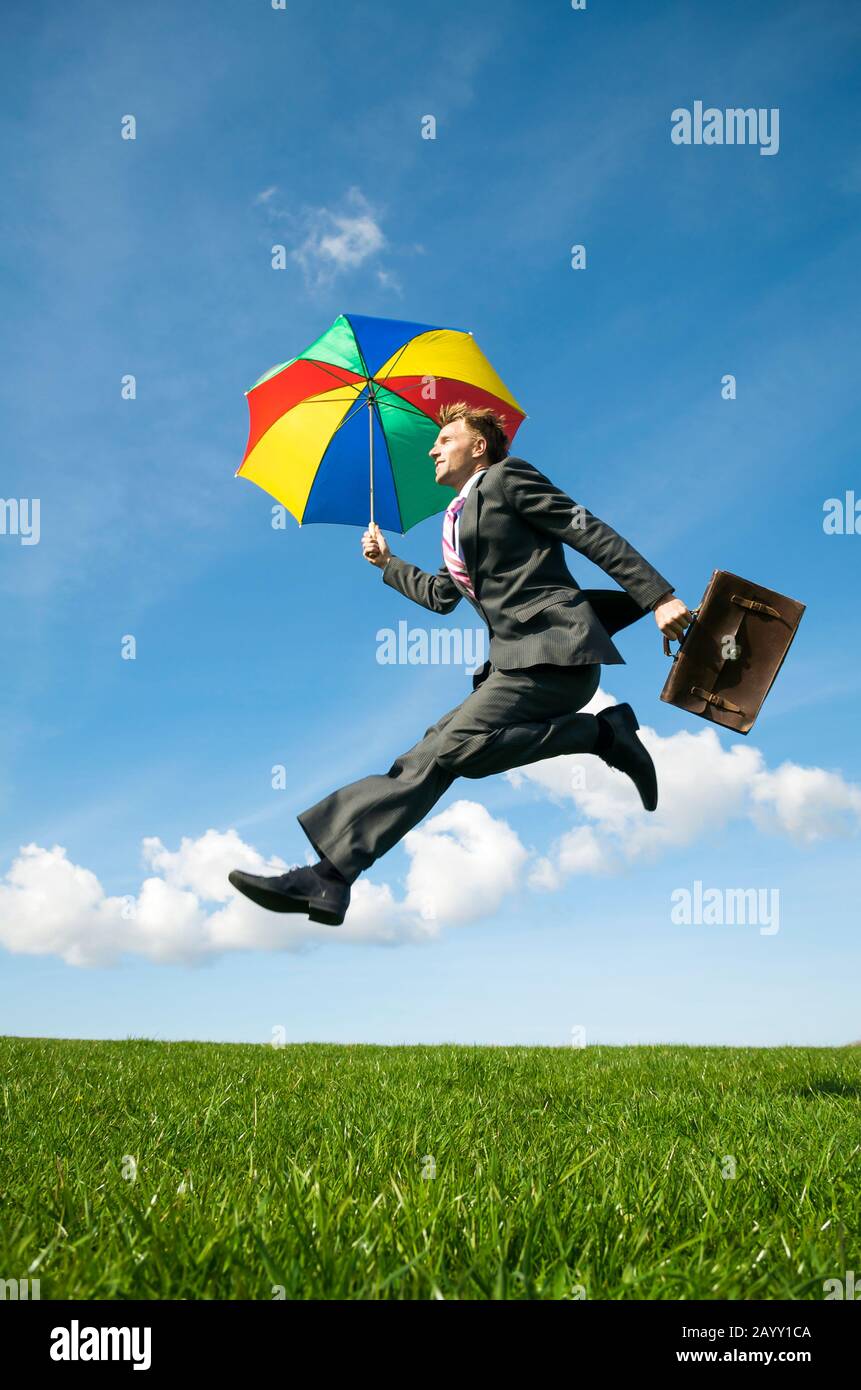 Lavoratore di ufficio con ombrello colorato e valigetta salto all'aperto attraverso campo di erba verde Foto Stock