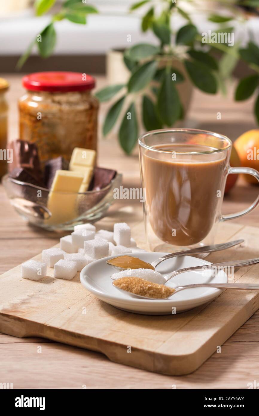 Lo zucchero in pezzatura, lo zucchero di canna, lo zucchero bianco sciolto e il miele di grano saraceno su un cucchiaino si trova accanto a un bicchiere con caffè e latte. In background cioccolato e. Foto Stock