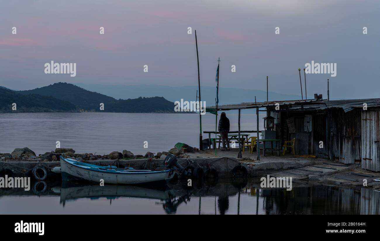 Zepko, Grecia - 26 aprile 2019: Piccolo porto al tramonto con piccola barca a remi e cabina con sedie e tavolo e sullo sfondo alcune colline e rosso Foto Stock