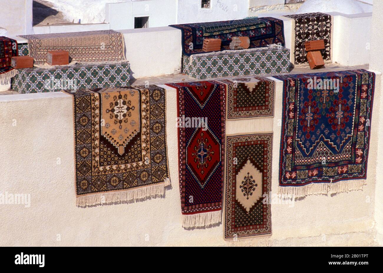 Tunisia: Tappeti orientali che si asciugano al sole sulle pareti bianche della medina Hammamet. Hammamet (dall'arabo al-hammamat, 'le terme') è conosciuta fin dall'epoca romana per la proprietà curativa delle sue acque. Le rovine romane sopravvivono ancora nei siti vicini di Siagum e Pupput, ma Hammamet nella sua forma attuale risale alla metà del XV secolo, quando furono costruiti la città vecchia, nota come medina, e il suq, o bazar principale. Hammamet medievale era preparata per la difesa - gli spagnoli fortificarono la kasbah nel XVI secolo - ma in realtà la storia della città è stata piacevolmente pacifica. Foto Stock
