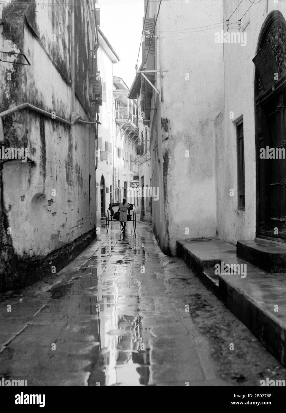 Tanzania/Zanzibar: Un estrattore di risciò su una strada di Stone Town dopo la pioggia, 1936. Zanzibar è una parte semi-autonoma della Tanzania nell'Africa orientale. È composto dall'arcipelago di Zanzibar nell'Oceano Indiano, 25-50 km (16-31 miglia) al largo della costa continentale, ed è costituito da numerose piccole isole e due grandi: Unguja (l'isola principale, indicata informalmente come Zanzibar), e Pemba. La capitale di Zanzibar, situata sull'isola di Unguja, è Zanzibar City. Il suo centro storico, conosciuto come Stone Town, è un sito Patrimonio dell'Umanità. Foto Stock