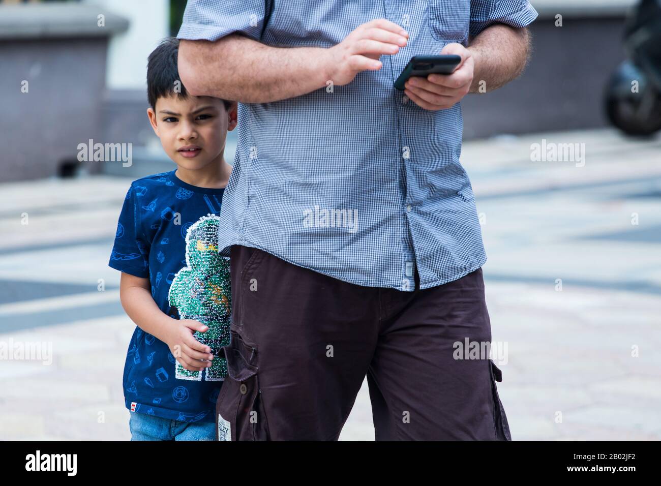 Un giovane e timido ragazzo arabico cerca di nascondersi dietro il padre mentre l'uomo o probabilmente suo padre è occupato sul suo cellulare. Foto Stock