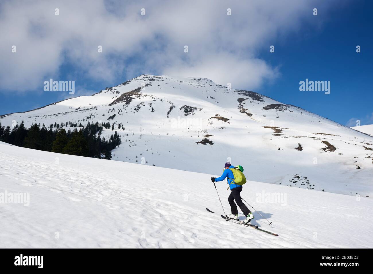 Escursionista maschile, turista in abiti colorati con zaino escursioni sugli sci su collina innevata sullo sfondo di cielo blu brillante e bella montagna in gelata giornata di sole. Concetto di stile di vita attivo invernale. Foto Stock