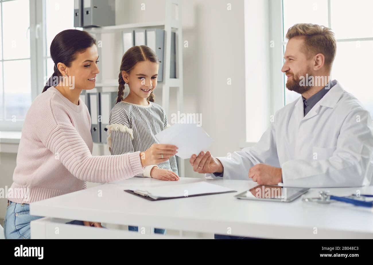 Il medico maschile dà a mamma e figlia una prescrizione mentre si siede a un tavolo in un ufficio clinico. Foto Stock