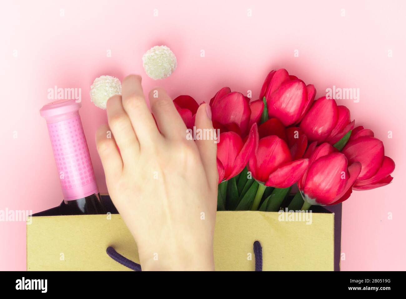 Concetto del regalo con vino e tulipani rossi nel sacchetto di carta sullo sfondo rosa. Disposizione piatta, vista dall'alto, spazio di copia. La mano della donna prende i dolci dal Foto Stock