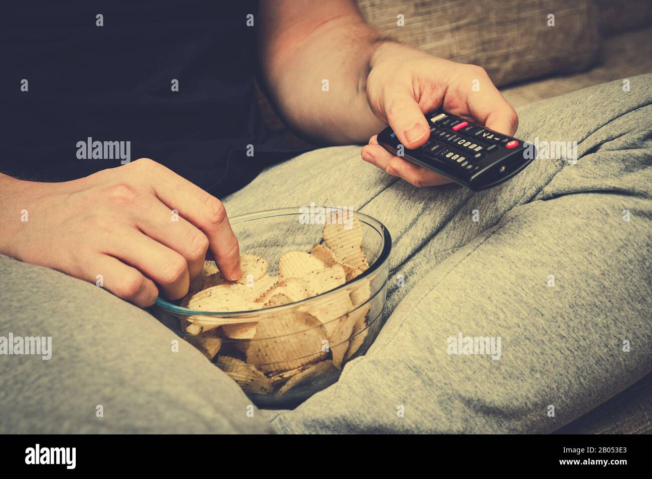 Un uomo si siede su un divano che mangia patatine dalla vetreria e cambia i canali con un telecomando TV. Tonato. Primo piano. Foto Stock