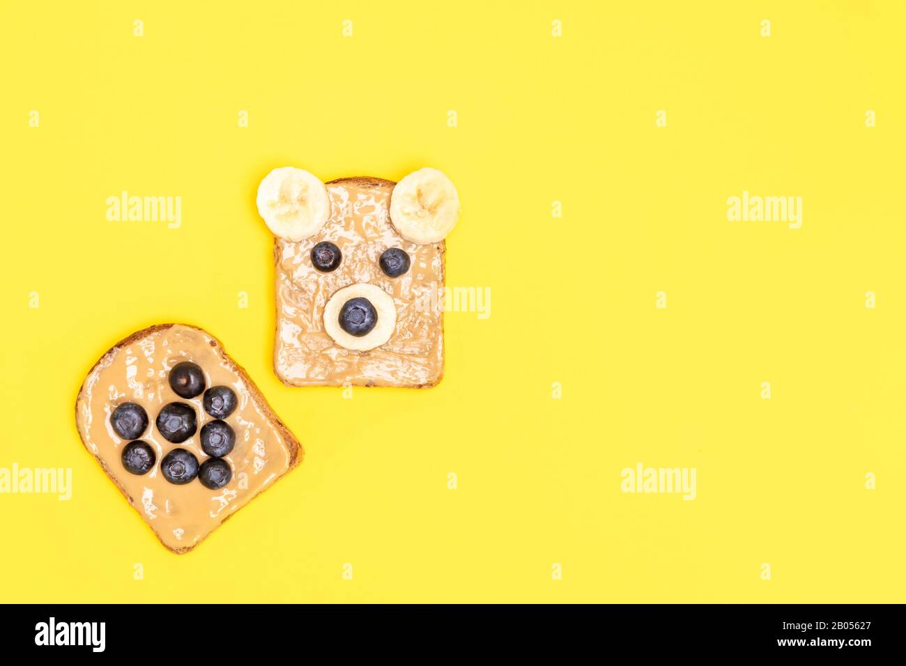 Divertenti arrosti di burro di arachide con la forma del cuore e il viso divertente per i bambini colazione su sfondo giallo. Vista dall'alto con spazio di copia Foto Stock
