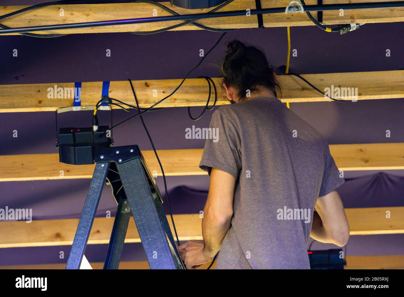 Una vista perdente e posteriore di un uomo al backstage di lavoro durante un festival multiculturale, lavoro a stagehand da scale che preparano attrezzature per musica set Foto Stock