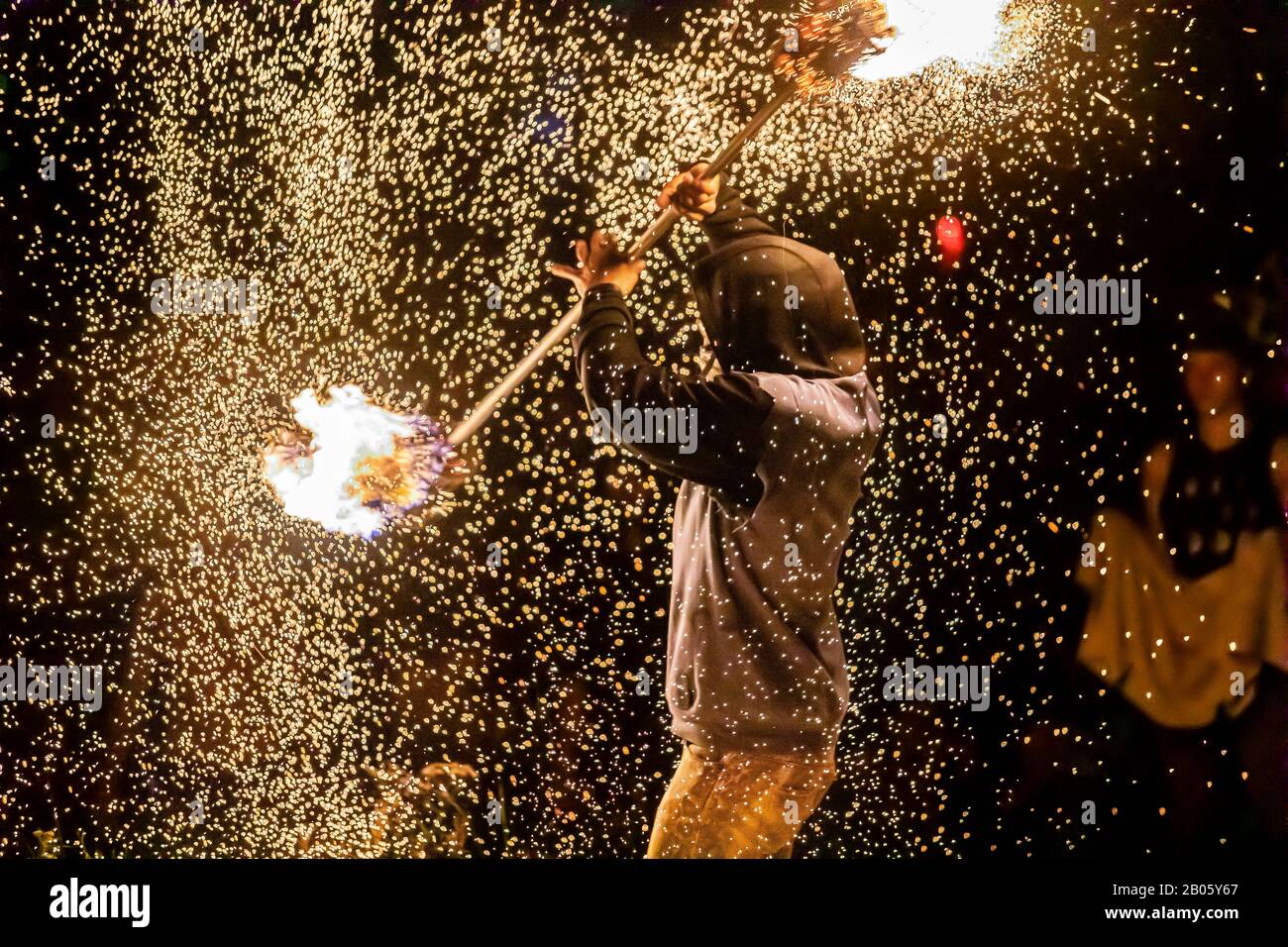 Un artista di danza del fuoco è visto usando una lunga lancia con fiamme accese ad ogni estremità, scintille calde e braci volare fuori durante la danza energetica all'aperto di notte Foto Stock