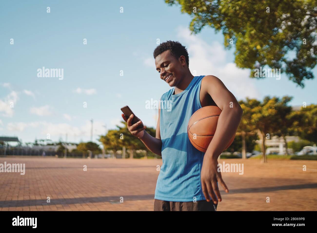 Felice giovane, sportivo giocatore di basket che tiene la palla in braccio utilizzando il telefono cellulare al parco Foto Stock